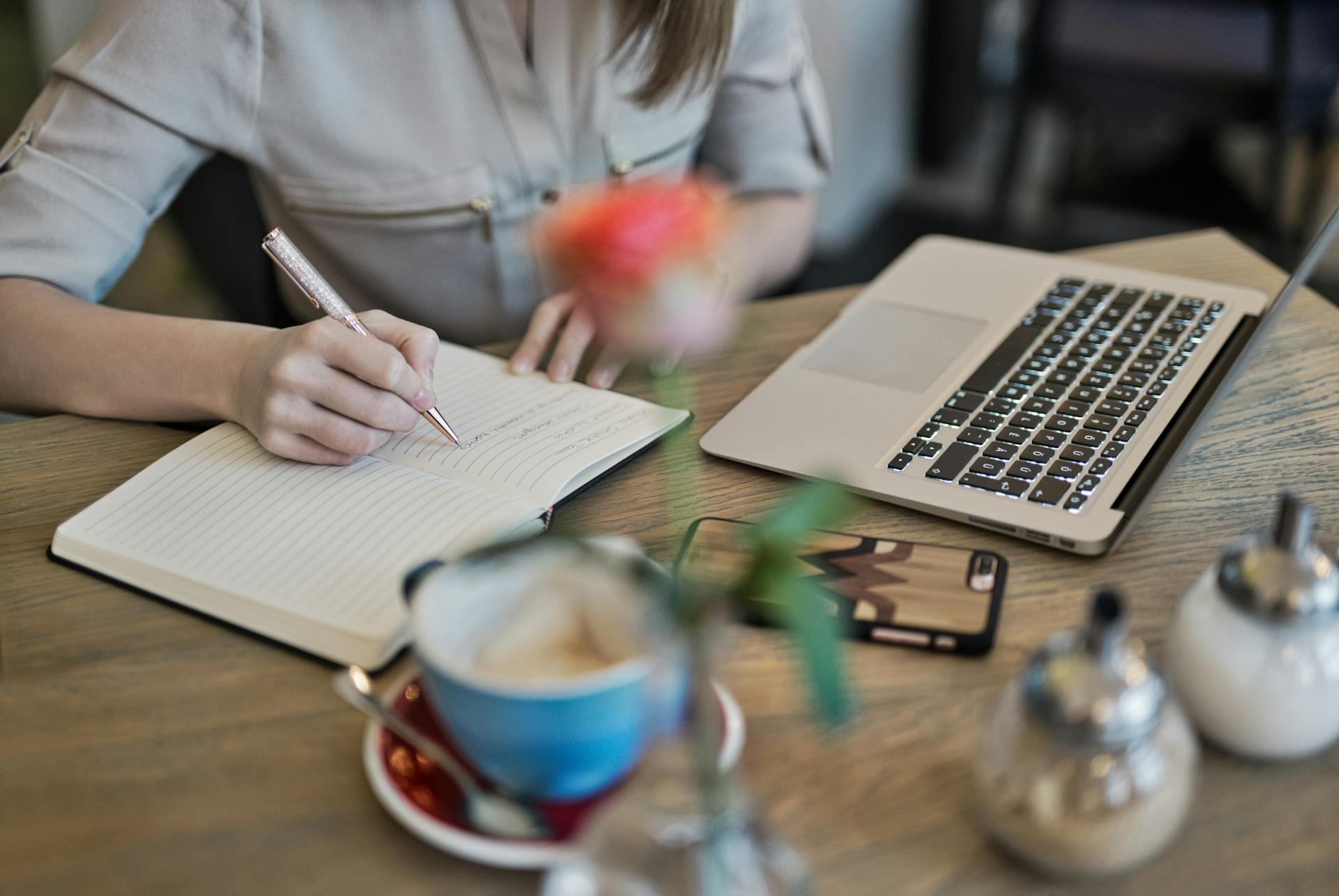 A hand holding a pen writing on a desk with a cup of coffee and a laptop next to it.