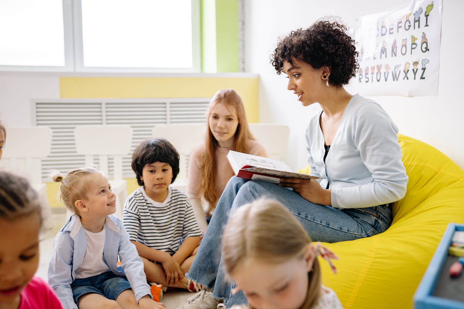 An adult, either a parent or a teacher, is holding a book and talking to three children, with another child in the same room.